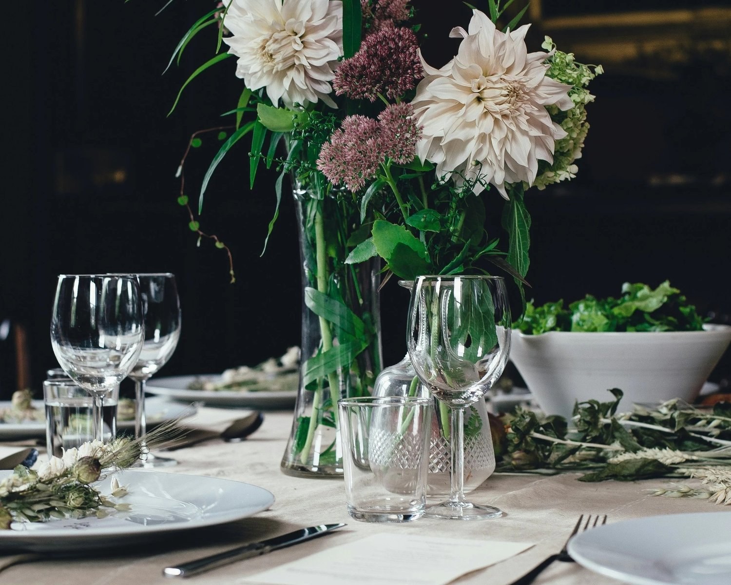 Dinner table set with white dahlias, greenery, crystal glassware and linen in soft candlelight