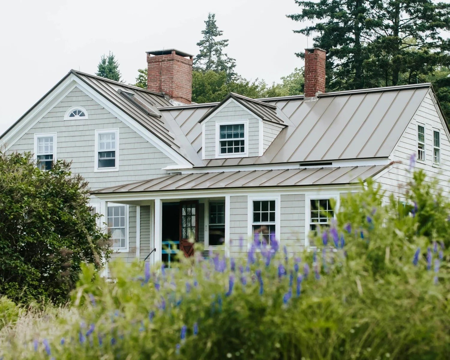 Shingled shore home with metal roofs and brick chimneys behind a lavender garden